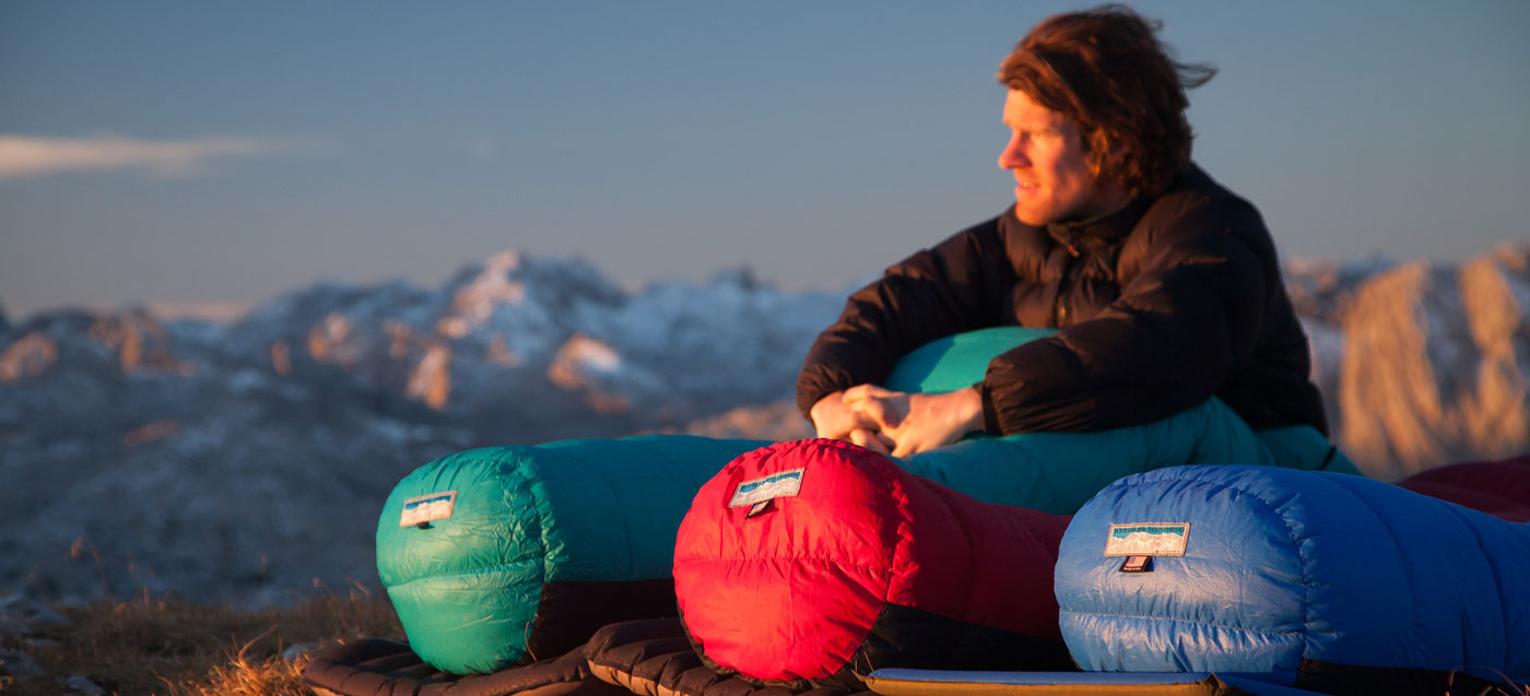 A person sits up in a sleeping bag and watches the sunset while camping in the mountains