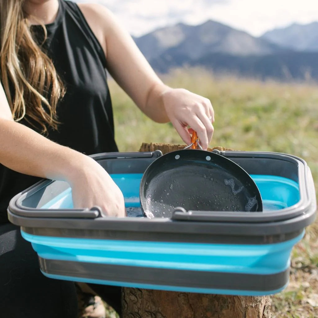 Person washing a pan in a blue collapsible sink outdoors with mountains in the background